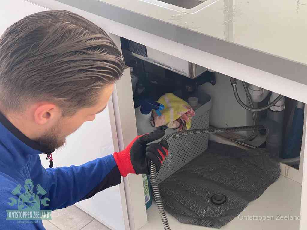 Een monteur voert een flexibele ontstoppingsveer in onder een keukenkastje om een verstopping voorbij de sifon te bereiken in Zeeland