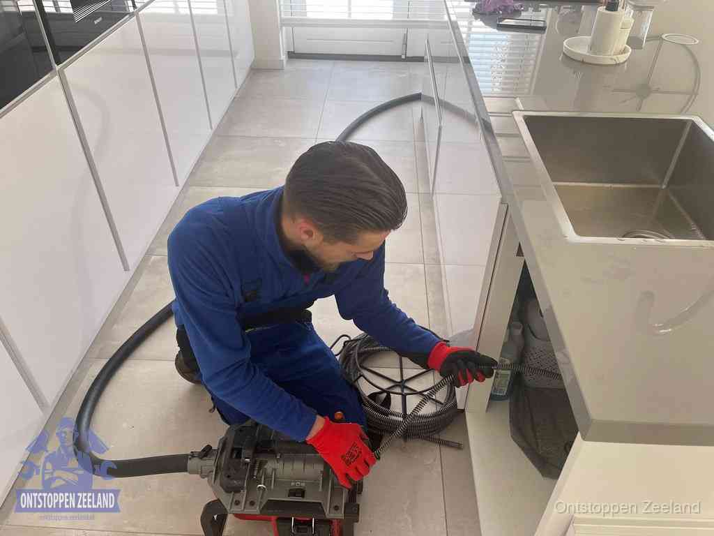 Een monteur ontstopt de keukenafvoer onder de spoelbak met een elektrische veermachine in Zeeland