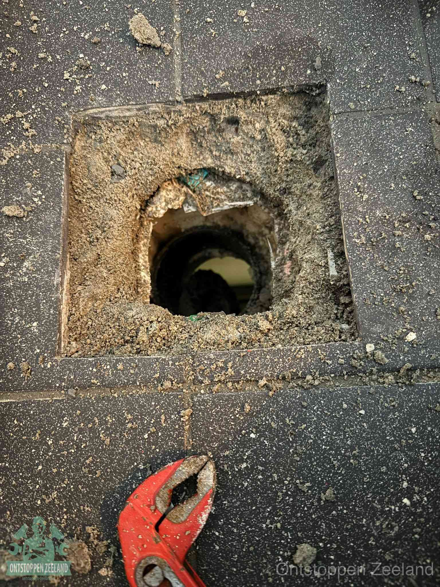 Een geopend rioolputje in een betontegelvloer met losgemaakt rooster en een zichtbare ronde afvoerbuis, het achterhalen waar de afvoerverstopping zit in Zeeland