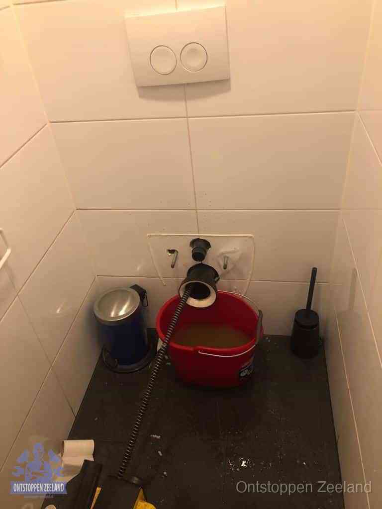 Een ontstopte wand-wc met verwijderde toiletpot waar een ontstoppingsveer in de wanduitgang verdwijnt, het achterhalen waar de verstopping zit in Zeeland