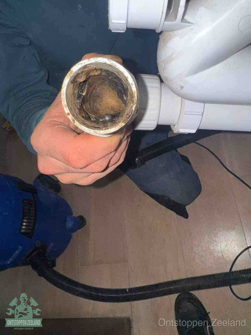 Een hand houdt een losgekoppeld PVC-afvoerbuisstuk vast dat van binnen volledig is dichtgeslibd met vet, vuil en aanslag, de meest voorkomende oorzaak van een verstopte afvoer in Zeeland