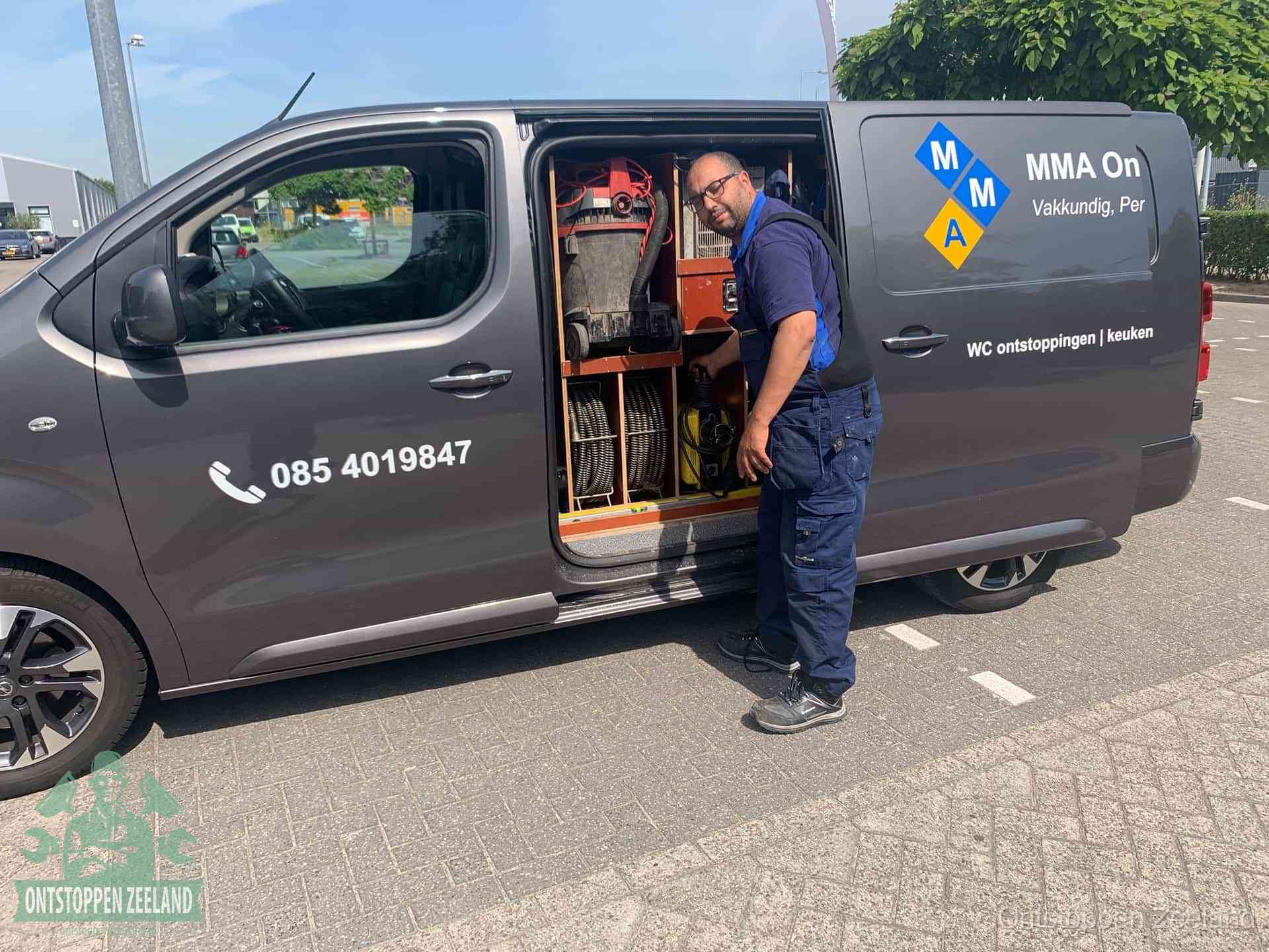 Een monteur bij de open schuifdeur van zijn bedrijfsbus op een parkeerplaats, klaar om de oorzaak van terugkerende afvoerverstoppingen aan te pakken in Zeeland