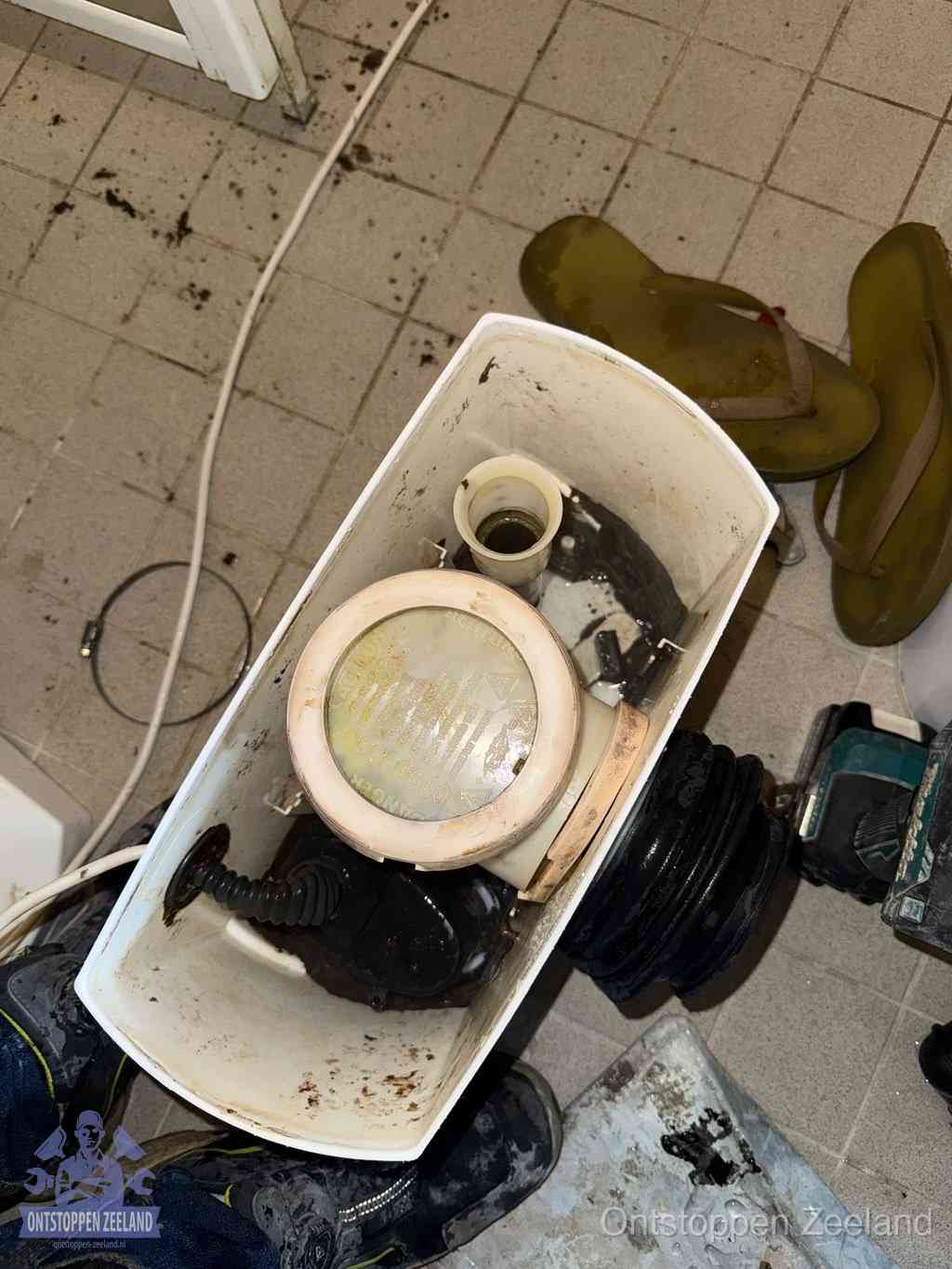 Een gedemonteerde toiletpot op de vloer met ontstoppingsveer en gereedschap ernaast, het verwijderen van een vastzittend voorwerp uit de wc in Zeeland