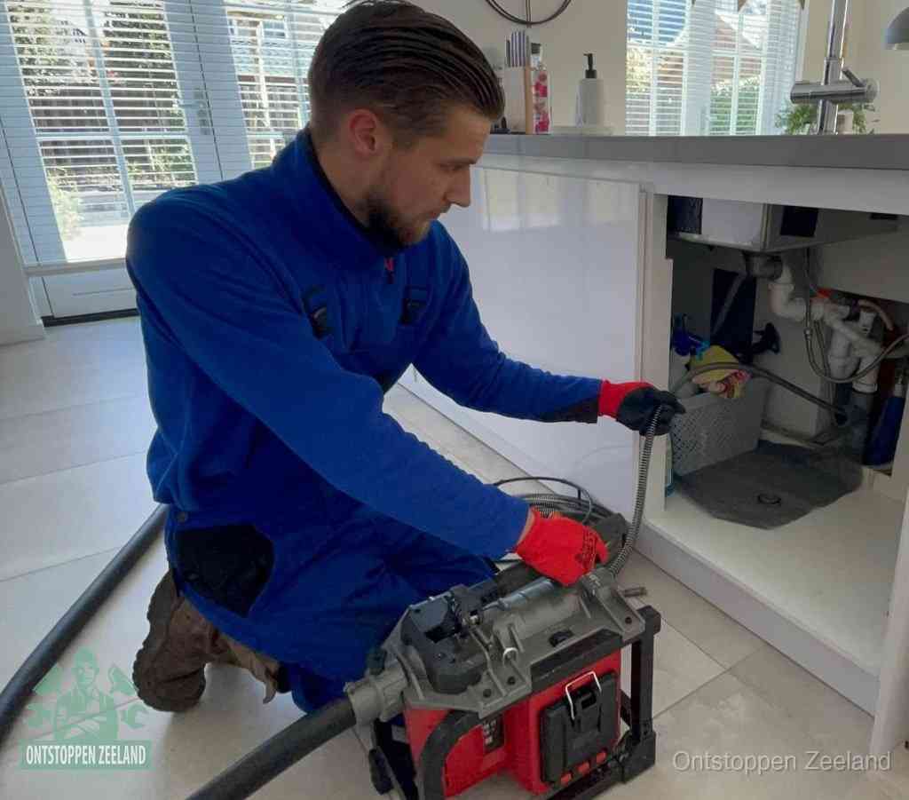 Een monteur met rode handschoenen knielt in een keuken en voert een elektrische ontstoppingsveer in tijdens het professioneel afvoer ontstoppen in Zeeland